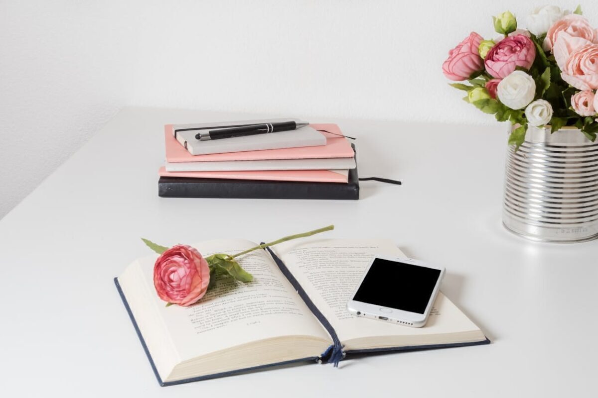 A stylish home office setup with open book, smartphone, and flowers on a white desk.